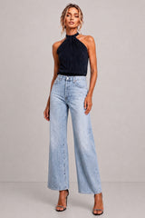 A model wearing a dark navy halter-neck top and light-wash wide-leg denim jeans, standing against a neutral gray studio background.