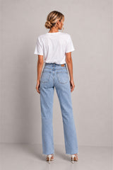 A woman viewed from the back wearing light-wash blue straight-leg jeans, a white short-sleeve t-shirt, and white heeled sandals against a plain light gray studio background.