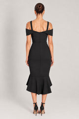 A woman viewed from the back wearing a black cold-shoulder midi dress with a fitted silhouette and ruffled hem, paired with black high-heeled sandals, against a light gray studio background.