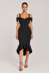 A woman modeling a black off-the-shoulder midi dress with a fitted silhouette and ruffled hem, paired with black strappy high-heeled sandals, against a light gray studio background.