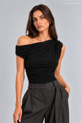A woman modeling a black off-the-shoulder ruched top paired with dark gray wide-leg trousers, photographed against a neutral light gray studio background.