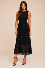 A woman modeling a sleeveless black midi dress with a pleated skirt and sheer tulle hem, paired with black high-heeled sandals, against a solid beige studio background.