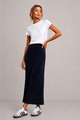 A woman modeling a white short-sleeve crewneck t-shirt and a dark navy blue pleated midi skirt, paired with white sneakers, against a neutral gray studio background.