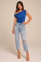 A woman standing wearing a blue draped one-shoulder top and light-wash blue jeans, standing against a plain beige studio background.
