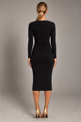A woman viewed from the back wearing a fitted black long-sleeve midi dress with gathered detailing at the waist, paired with metallic gold high-heeled sandals, against a neutral gray studio background.