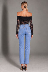 A woman shown from the back wearing a black off-the-shoulder lace long-sleeve top and light blue straight-leg jeans, standing against a gray studio background.