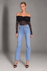A model wearing a black off-the-shoulder lace long-sleeve top and light-wash blue straight-leg jeans, standing against a neutral gray studio background.