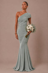A woman models a one-shoulder floor-length gown in a muted sage green color, holding a bouquet of white flowers against a solid taupe background.