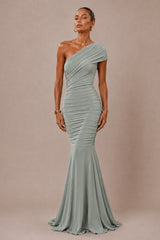 A woman modeling a one-shoulder floor-length gown in a dusty sage green color, set against a solid warm taupe background.