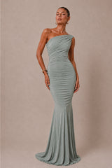 A woman modeling a one-shoulder ruched mermaid gown in a dusty sage green color, set against a solid beige studio backdrop.