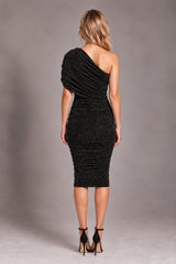 A woman viewed from the back wearing a black one-shoulder ruched bodycon dress with black high heels, standing against a neutral gray studio background.