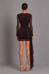 A woman viewed from the back wearing a dark brown long-sleeve midi dress with sheer mesh sleeves and a ruched waist, paired with metallic gold high-heeled sandals, against a solid gray studio background.