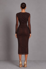 A woman viewed from the back wearing a dark brown long-sleeve midi dress with sheer mesh sleeves and a ruched waist, paired with metallic gold high-heeled sandals, against a solid gray studio background.