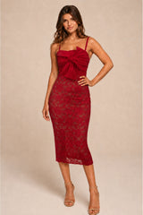 A woman modeling a deep red lace midi dress with spaghetti straps, a sweetheart neckline, and a large gathered bow detail at the bust, posed against a solid beige studio backdrop.
