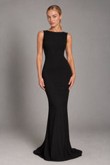 A woman modeling a sleeveless, floor-length black gown with a boat neckline and a slight train, photographed against a plain gray studio background.