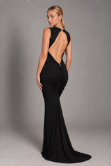 A woman modeling a floor-length black evening gown from the back, featuring a dramatic open back, ruched detailing, and a slight train, set against a plain gray studio background.