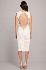 A woman viewed from the back wearing a fitted sleeveless dress with a large open back and ruched texture, paired with nude high heels, against a light gray studio background.