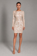 A woman modeling a fitted long-sleeve mini dress covered in beige and cream floral lace, paired with clear high-heeled sandals, against a solid gray studio background.
