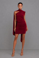 A model wearing a deep burgundy berry one-shoulder mini dress with a draped cape detail, paired with gold high-heeled sandals, set against a solid grey studio background.
