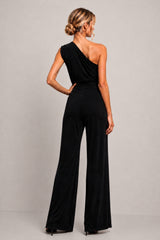 A woman modeling the back of a black one-shoulder wide-leg jumpsuit against a neutral light gray studio background.