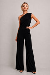 A woman modeling a black one-shoulder wide-leg jumpsuit against a neutral light gray studio background.