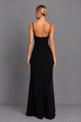 A woman viewed from the back wearing a floor-length black gown with thin spaghetti straps and a low open back, standing against a plain gray studio backdrop.