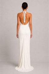 A woman viewed from the back wearing a floor-length white halter gown with an open back and gathered skirt, standing against a plain light gray studio background.