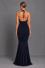 A woman viewed from the back wearing a sleeveless, open-back mermaid gown in dark navy blue, standing against a plain gray studio backdrop.