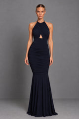 A woman modeling a sleeveless, high-neck, floor-length navy blue gown with a ruched bodice and a small triangular cutout at the chest, photographed against a plain gray studio backdrop.
