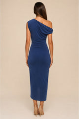 A woman viewed from the back wearing a fitted one-shoulder midi dress in a medium-to-deep blue color, paired with beige high-heeled sandals, against a solid light beige studio background.