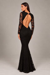A woman modeling a black lace mermaid gown from the back, featuring a dramatic open back, long lace sleeves, and a fishtailed lace-trimmed hem, set against a plain gray studio background.