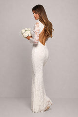 A woman modeling a fitted white lace wedding gown with long sheer lace sleeves, a low open back, and a mermaid silhouette, standing against a light gray studio background and holding a bouquet of white flowers.