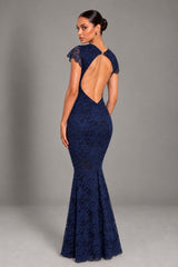 A woman modeling a navy blue lace mermaid gown from the back, featuring a dramatic open back, short cap sleeves, and a fitted silhouette, set against a neutral gray studio background.