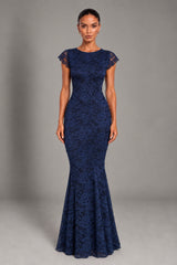 A woman modeling a navy blue lace mermaid gown with cap sleeves against a neutral gray studio background.