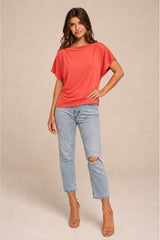 A woman modeling a coral orange short-sleeve knit top with light-wash distressed jeans and tan heeled sandals, standing against a plain beige studio background.