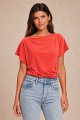 A woman modeling a bright coral-red short-sleeve top with a wide boat neckline, paired with light-wash blue jeans, against a plain beige studio background.
