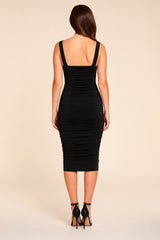 A woman viewed from the back wearing a black ruched midi dress with a square neckline and shoulder straps, paired with black high-heeled sandals, against a solid beige studio background.