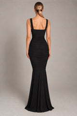 A woman viewed from the back wearing a fitted black sleeveless gown with a square neckline, ruched bodice, and flared mermaid-style hem, photographed against a plain light gray studio background.