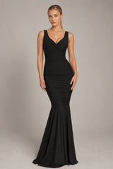A woman modeling a sleeveless black mermaid gown with a deep V-neckline and ruched bodice, photographed against a plain light gray studio background.