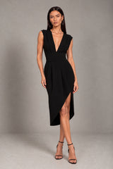 A woman modeling a sleeveless black midi dress with a deep V-neckline, defined waist seam, and asymmetrical high-low hem with a front slit, paired with black strappy heels against a neutral gray studio backdrop.
