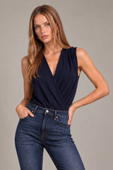 A woman modeling a sleeveless navy wrap-style top with a deep V-neckline, paired with dark blue jeans, against a solid gray studio background.