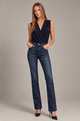 A woman modeling dark blue straight-leg jeans with a navy sleeveless wrap top and black heels, standing against a neutral gray studio background.