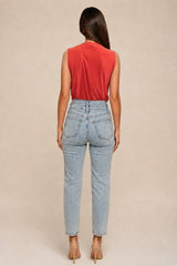 A woman shown from the back wearing a sleeveless red top and light-wash blue jeans, standing against a plain beige studio background.