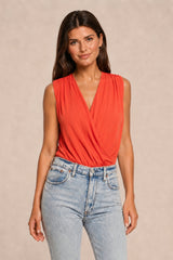 A woman modeling a sleeveless coral-orange wrap-style top paired with light-wash blue jeans, standing against a plain beige studio background.
