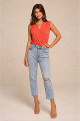 A woman modeling a sleeveless coral wrap-style top and light-wash distressed cropped jeans with tan heeled sandals, standing against a plain beige studio background.