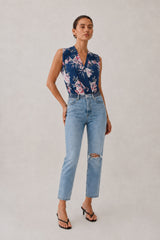 A woman modeling a sleeveless navy floral top and light-wash distressed jeans with black thong sandals, standing against a plain light gray studio background.