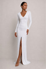 A woman modeling a fitted white long-sleeve maxi dress with a deep V-neckline, ruched waist, and a high front slit, photographed against a plain light beige studio backdrop.