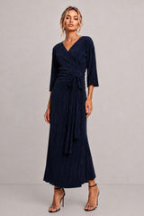A woman modeling a navy blue pleated wrap maxi dress with three-quarter sleeves and a self-tie waist, paired with black high-heeled sandals against a neutral gray studio background.