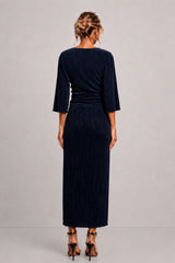 A woman viewed from the back wearing a dark navy pleated maxi dress with three-quarter sleeves and black high-heeled sandals, standing against a light gray studio background.
