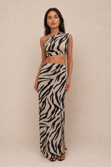 A woman modeling a two-piece zebra-print outfit consisting of a one-shoulder crop top and a matching maxi skirt, set against a plain beige studio background.